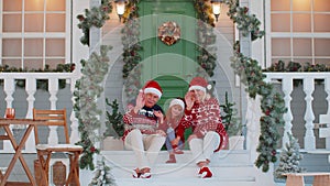 Senior grandmother grandfather with granddaughter sitting at Christmas house porch waving hello hi