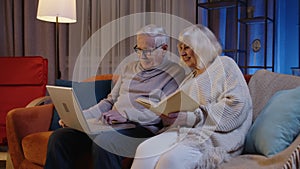 Senior grandparents couple reading book, using laptop pc on couch in night living room at home