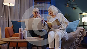 Senior grandparents couple reading book, using laptop pc on couch in night living room at home