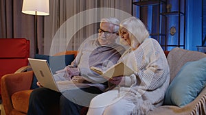 Senior grandparents couple reading book, using laptop pc on couch in night living room at home