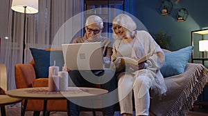 Senior grandparents couple reading book, using laptop pc on couch in night living room at home