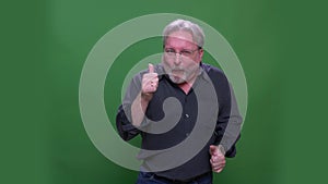 Senior gray haired american manin glasses dances and shows thumbs-up isolated on green chromakey background.