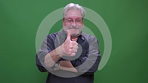 Senior gray haired american manin glasses puts thumb-up to show respect isolated on green chromakey background.