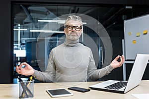Senior gray haired businessman meditating inside office building sitting on chair at desk, mature man wearing glasses