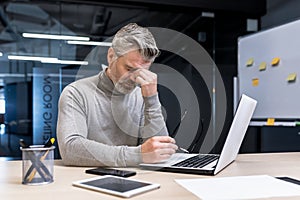 Senior gray haired businessman overtired working on laptop, mature man wearing glasses has pain in eyes, boss working in photo