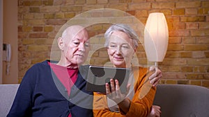 Senior gray-haired caucasian couple having online videocall on tablet being happy and joyful at cozy home.