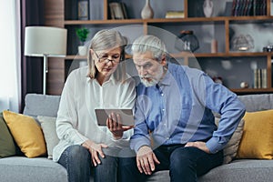 Senior gray-haired couple man and woman sitting on sofa at home. use a tablet computer for video communication and online