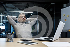 Senior gray haired man resting in office on chair with hands behind head, businessman boss working using laptop, mature