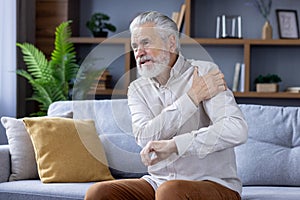 Senior gray-haired man sitting on sofa at home and holding arm above shoulder, suffering from pain and sprain