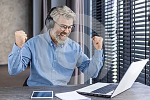Senior gray haired man wearing headphones is watching sports match at workplace inside office, joyful adult boss smiling