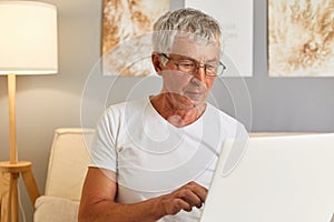 Senior gray haired man wearing white T-shier and glasses sitting on couch in living room holding laptop typing on keyboard looking