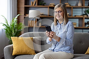 Senior gray-haired woman sitting joyfully on sofa at home, pensioner holding phone in hands, using app on smartphone