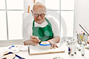 Senior grey-haired artist man smiling happy painting pottery sitting on the table at art studio