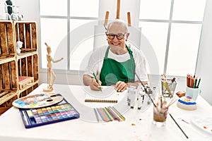 Senior grey-haired artist man smiling happy painting sitting on the table at art studio