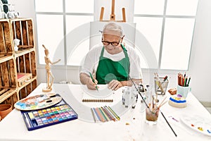 Senior grey-haired artist man smiling happy painting sitting on the table at art studio