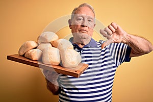 Senior grey haired baker man holding fresh homemade bread over yellow background with angry face, negative sign showing dislike