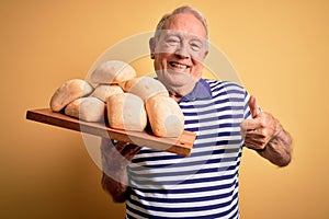 Senior grey haired baker man holding fresh homemade bread over yellow background happy with big smile doing ok sign, thumb up with