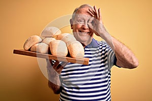Senior grey haired baker man holding fresh homemade bread over yellow background with happy face smiling doing ok sign with hand