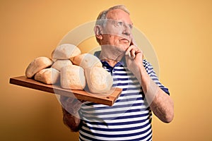Senior grey haired baker man holding fresh homemade bread over yellow background serious face thinking about question, very