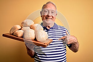 Senior grey haired baker man holding fresh homemade bread over yellow background with surprise face pointing finger to himself