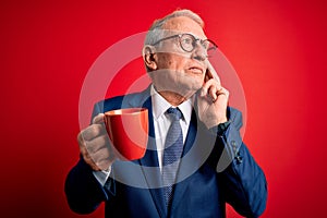 Senior grey haired business man drinking a hot cup of coffee over red background serious face thinking about question, very
