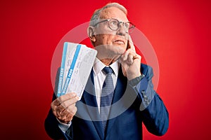 Senior grey haired business man holding airplane boarding pass over red background serious face thinking about question, very