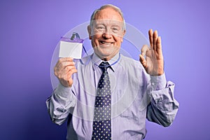 Senior grey haired business man holding identification tag over purple background doing ok sign with fingers, excellent symbol