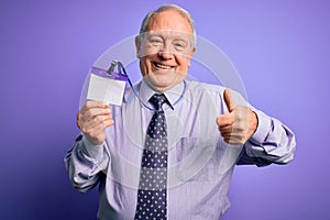 Senior grey haired business man holding identification tag over purple background happy with big smile doing ok sign, thumb up