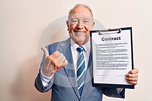 Senior grey-haired businessman wearing suit holding clipboard with contract document pointing thumb up to the side smiling happy