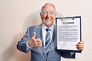 Senior grey-haired businessman wearing suit holding clipboard with contract document smiling happy and positive, thumb up doing