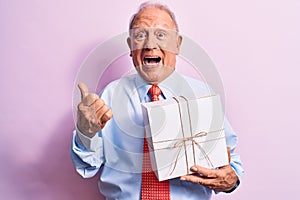 Senior grey-haired businessman wearing tie holding birthday gift over pink background pointing thumb up to the side smiling happy