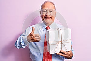Senior grey-haired businessman wearing tie holding birthday gift over pink background smiling happy and positive, thumb up doing