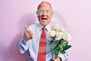 Senior grey-haired businessman wearing tie holding bouquet of flowers over pink background pointing thumb up to the side smiling