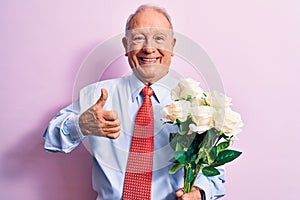Senior grey-haired businessman wearing tie holding bouquet of flowers over pink background smiling happy and positive, thumb up