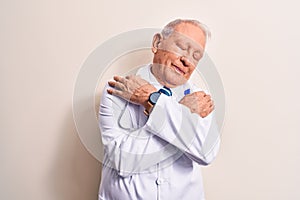 Senior grey-haired doctor man wearing coat and stethoscope standing over white background hugging oneself happy and positive,