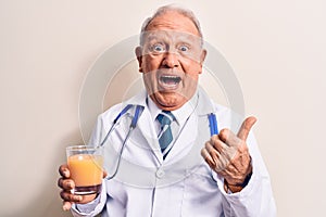 Senior grey-haired doctor man wearing stethoscope drinking glass of healthy orange juice pointing thumb up to the side smiling