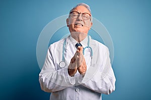 Senior grey haired doctor man wearing stethoscope and medical coat over blue background begging and praying with hands together