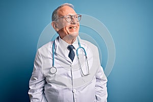 Senior grey haired doctor man wearing stethoscope and medical coat over blue background looking away to side with smile on face,