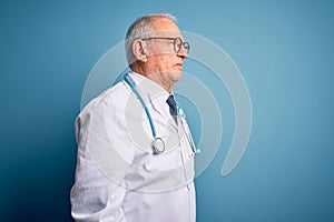 Senior grey haired doctor man wearing stethoscope and medical coat over blue background looking to side, relax profile pose with