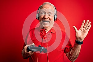 Senior grey haired gamer man playing video games using gamepad joystick over red background very happy and excited, winner