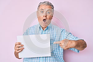 Senior grey-haired man holding blank empty banner afraid and shocked with surprise and amazed expression, fear and excited face