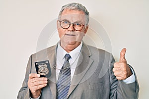 Senior grey-haired man holding detective badge smiling happy and positive, thumb up doing excellent and approval sign
