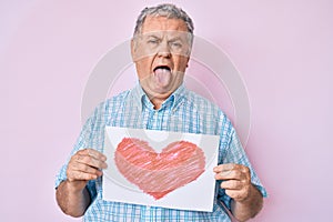 Senior grey-haired man holding heart draw sticking tongue out happy with funny expression