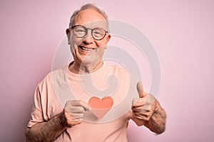 Senior grey haired man holding heart shape paper over pink background happy with big smile doing ok sign, thumb up with fingers,