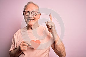 Senior grey haired man holding heart shape paper over pink background surprised with an idea or question pointing finger with