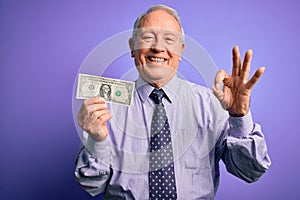 Senior grey haired man holding one dollar bank note over purple background doing ok sign with fingers, excellent symbol