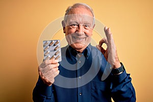 Senior grey haired man holding pharmaceutical pill drugs over yellow background doing ok sign with fingers, excellent symbol