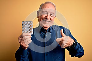 Senior grey haired man holding pharmaceutical pill drugs over yellow background happy with big smile doing ok sign, thumb up with