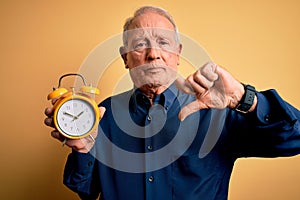 Senior grey haired man holding vintage alarm clock over yellow background with angry face, negative sign showing dislike with