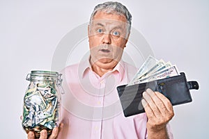 Senior grey-haired man holding wallet with dollars and jar with savings clueless and confused expression